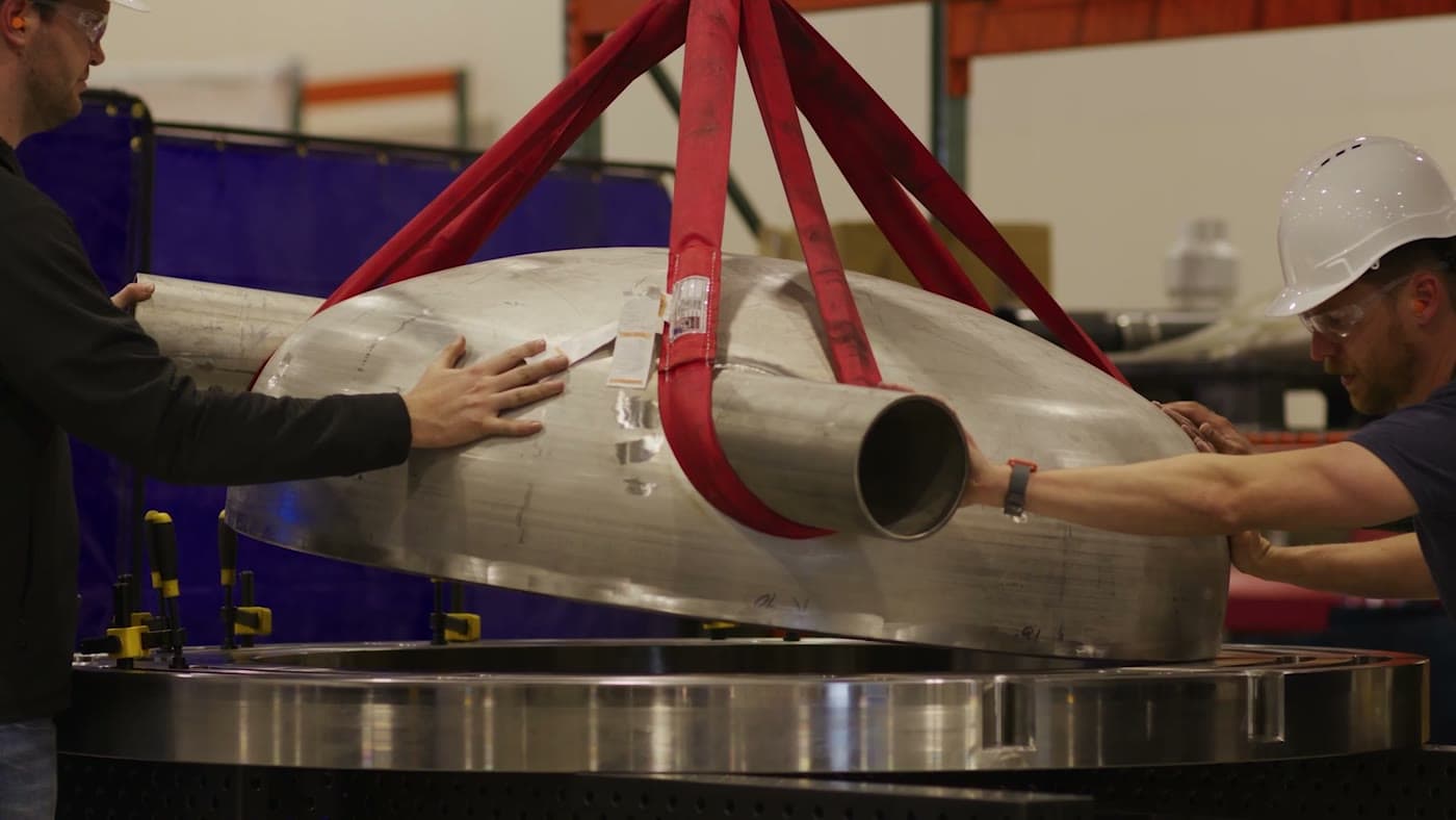 Hardware suspended in red lifting straps, guided by hand onto a test stand.